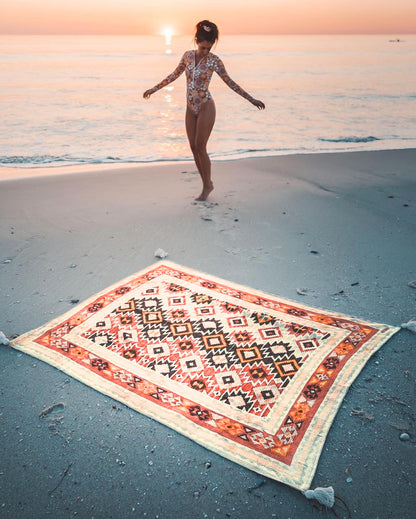 Person standing on a patterned rug on a beach at sunset